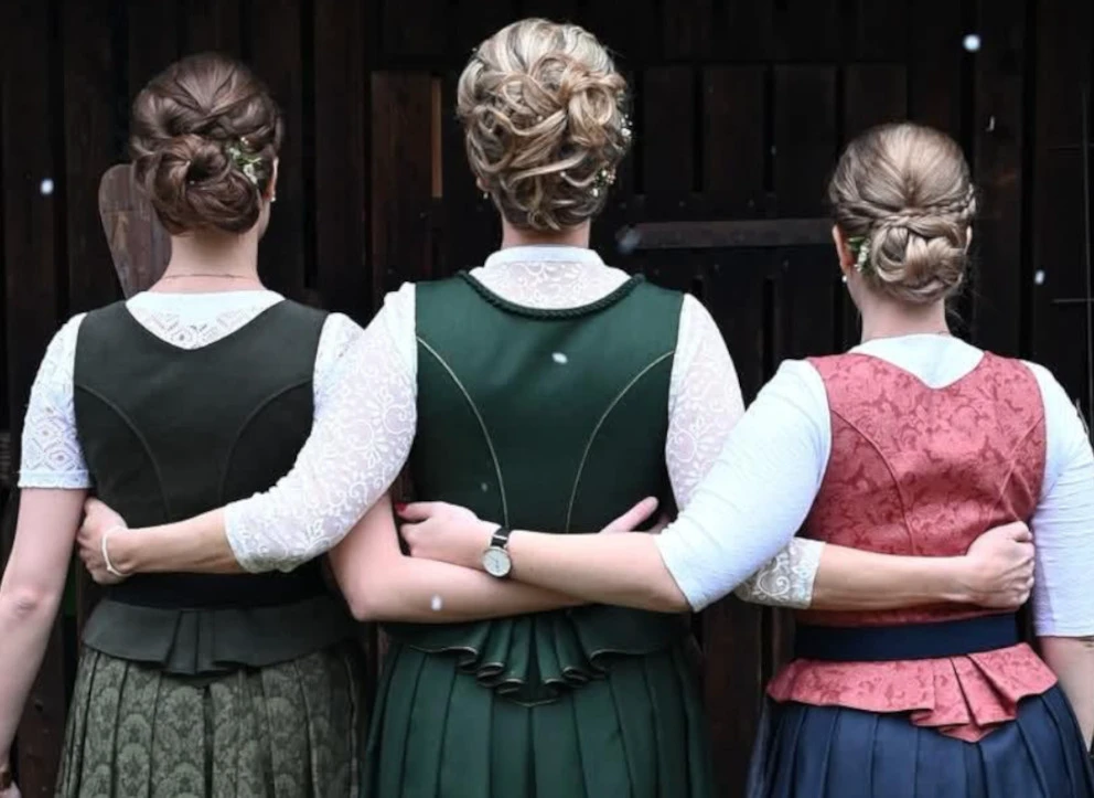 Drei Frauen in traditionelle Trachten umarmen sich vor einem Holzzaun