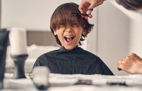 Ein fröhliches Kind mit offenem Mund während ein Friseur ihm die Haare schneidet