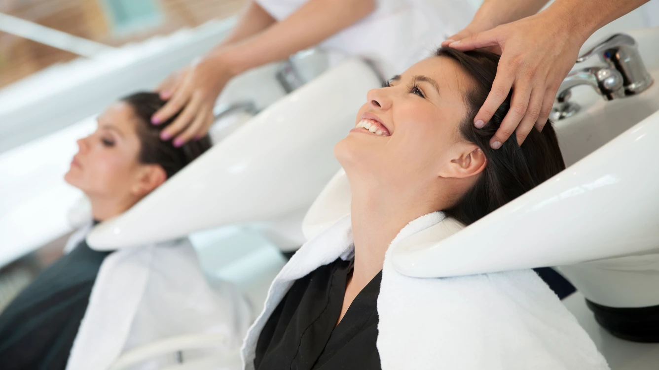 Eine Frau lächelt beim Haarwäschen in einem Salon während ein Stylist ihre Kopfhaut massiert